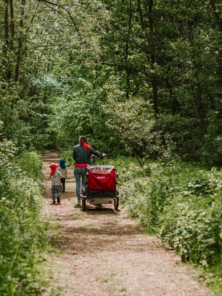 Frau schiebt Fahrrad mit Fahrradanhänger durch den Wald. Zwei Kinder laufen nebenher.